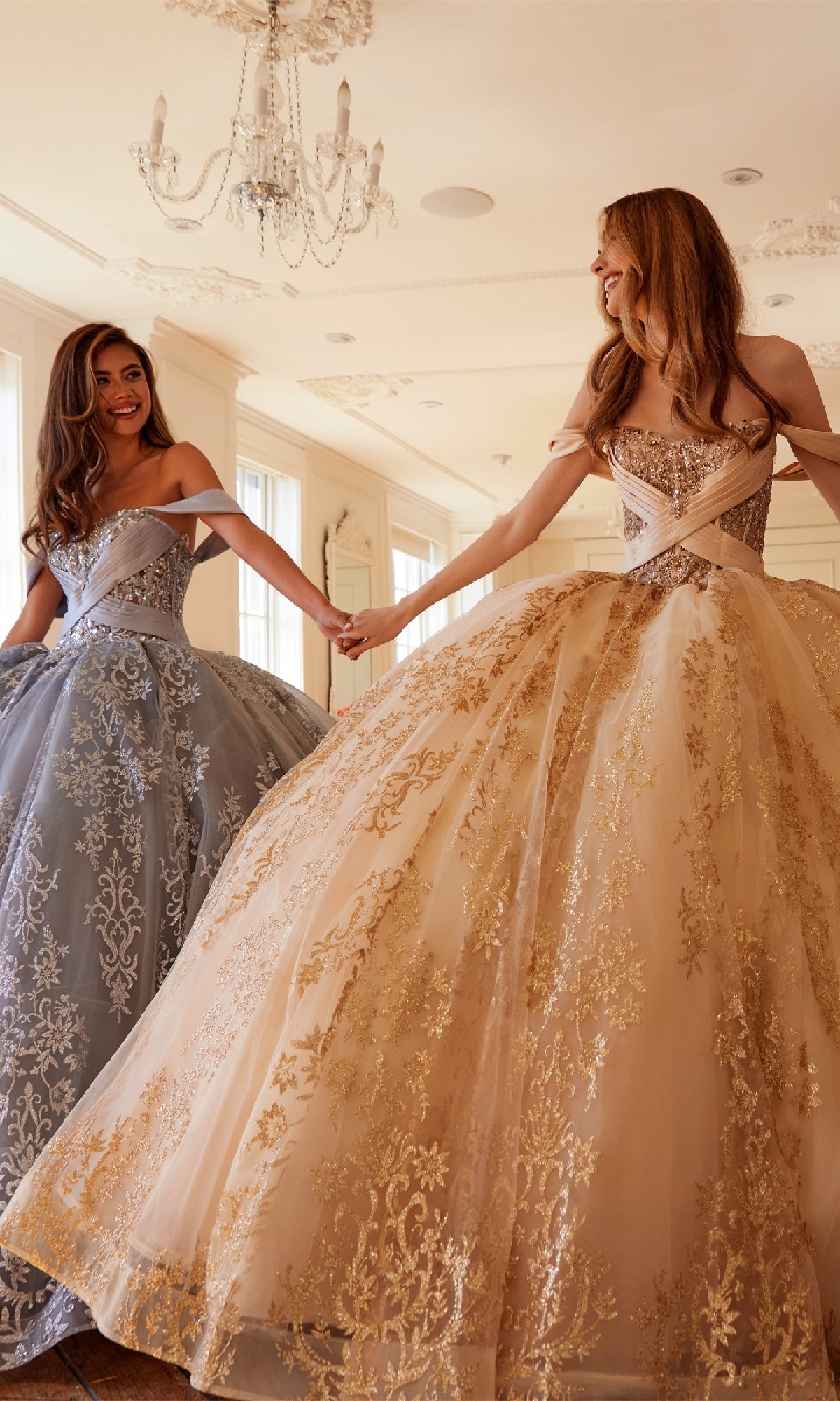 Two women smile and hold hands in a bright room with a chandelier, each wearing the Quinceanera Dress JT1488H By Juliet—ornate ball gowns featuring gold embroidery and corset bodices.